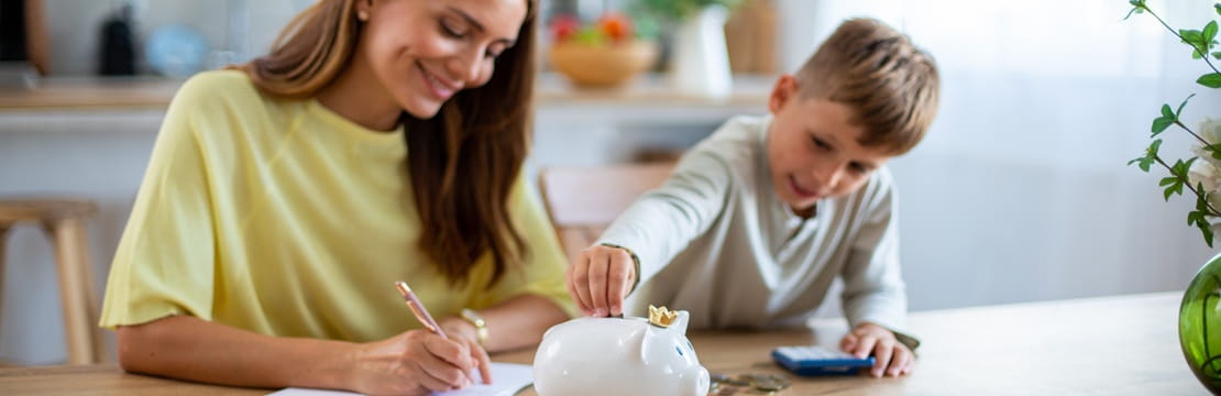 Mother and son sitting at a table, writing and placing coins into a white piggy bank.